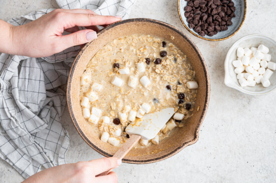 A woman's hand holding a silicone baking spatula mixing the s'mores baked oatmeal ingredients in a large tan bowl.