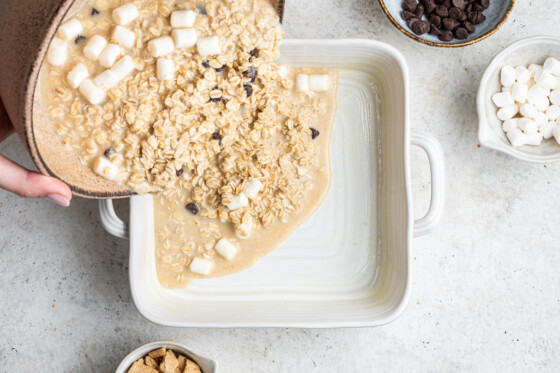 Woman's hands holding a large bowl containing the s'mores baked oatmeal ingredients. The ingredients are poured into a white square baking dish.