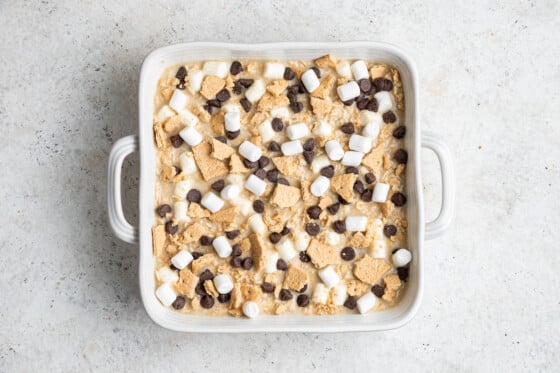 A white square baking dish with the s'mores baked oatmeal inside before baking in the oven. Topped with chocolate chips, mini marshmallows, and crushed graham crackers.