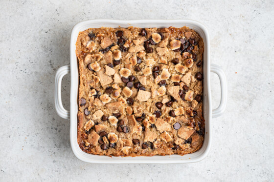 A white square baking dish with the s'mores baked oatmeal inside after baking in the oven. The top is lightly browned with chocolate chips, mini marshmallows, and crushed graham crackers.