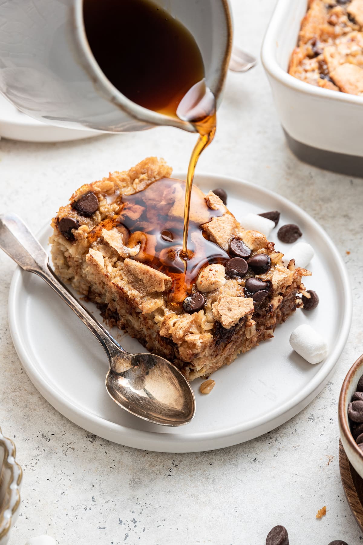 A white container of maple syrup is drizzled on top of a piece of the s'mores baked oatmeal placed on a small white plate with a spoon.