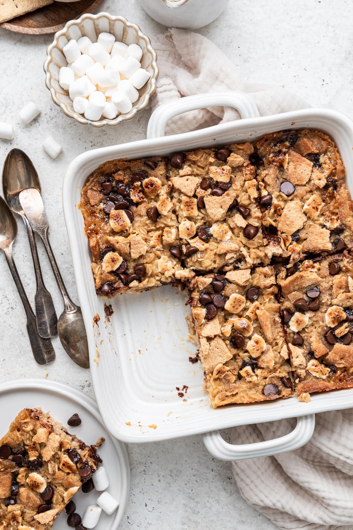 A square white baking dish containing five pieces of the s'mores baked oatmeal. One piece has been cut out of the baking dish and is on a small white plate nearby.