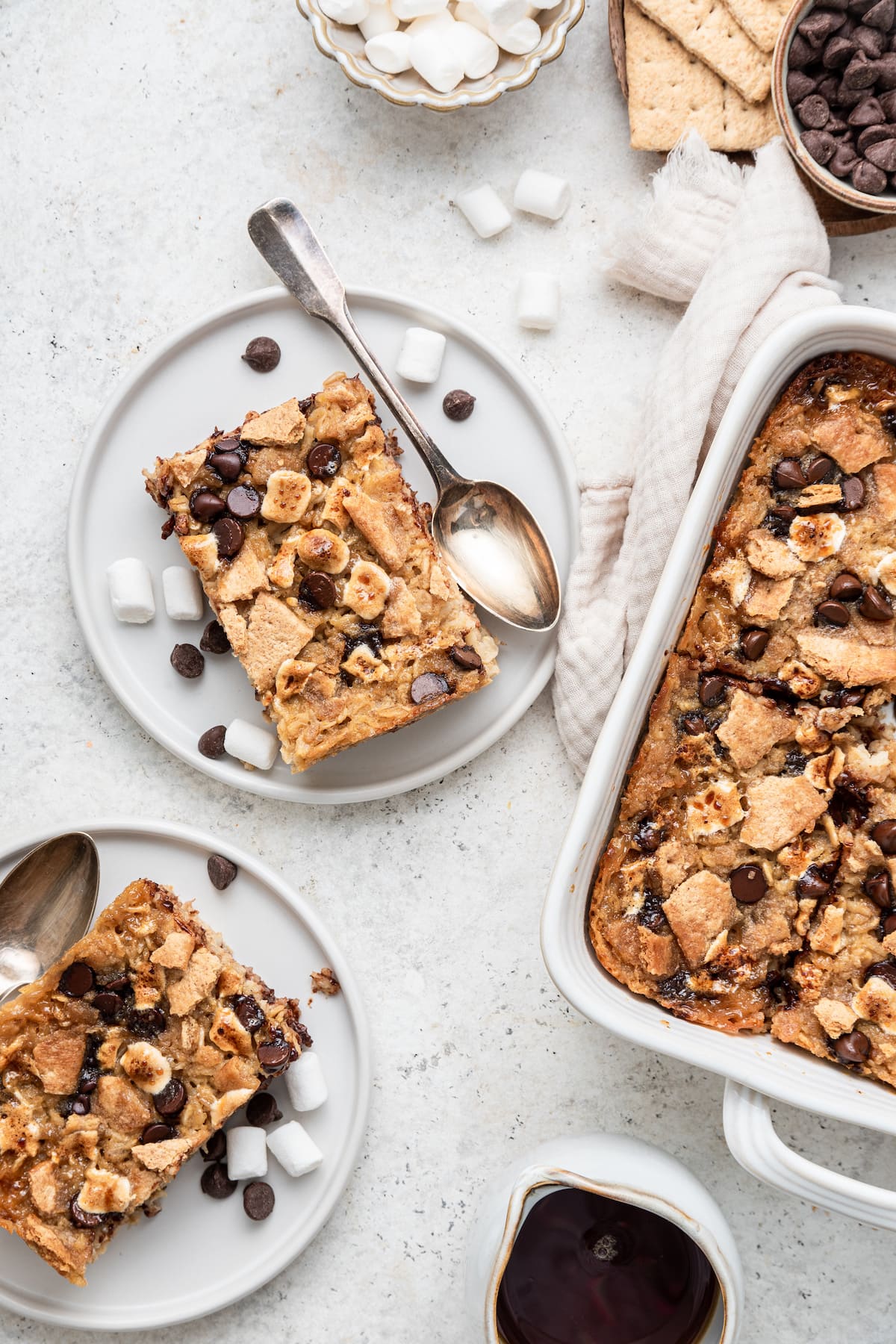 Two small white plates with spoons. On the plates are individual pieces of the s'mores baked oatmeal with mini marshmallows and chocolate chips.