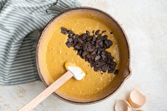 The tahini banana bread mixture as a liquid in a large tan bowl topped with chocolate chips with a silicon spatula inside.