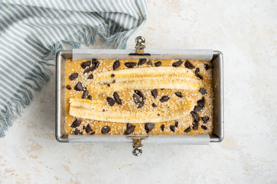 A top view of tahini banana bread inside a bread pan before being baked in the oven. The bread is topped with sesame seeds and chocolate chips.