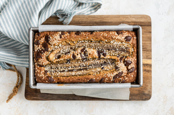 A top view of tahini banana bread inside a bread pan on a wooden cutting board after being baked in the oven. The bread is topped with sesame seeds and chocolate chips.