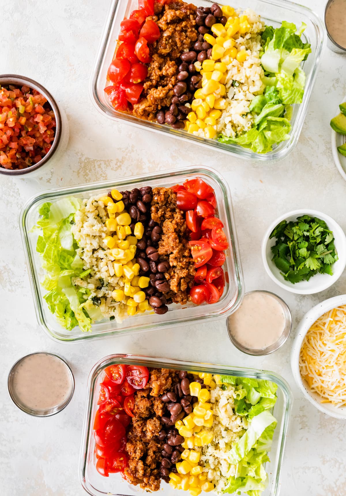 Three turkey taco bowls in glass meal prep containers, served with sides of dressing, cilantro, shredded cheese and pico de gallo.