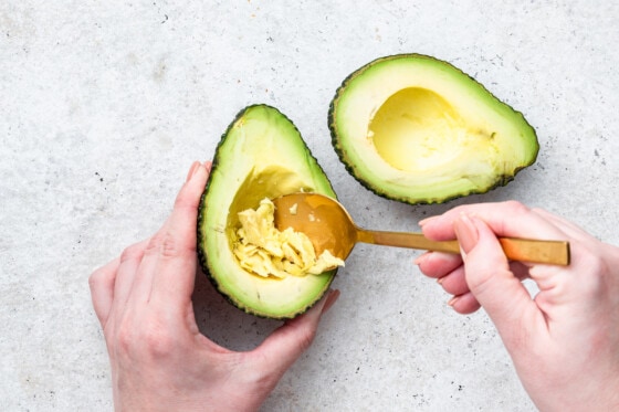 A woman's hand uses a golden spoon to scrap out some avocado to make a large hole.