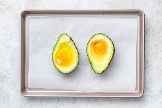 An avocado split in half with two raw eggs in the center of each half on a baking tray.