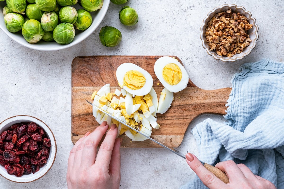 A woman's hand using a knife to chop two hard-boiled eggs on a wooden cutting board.