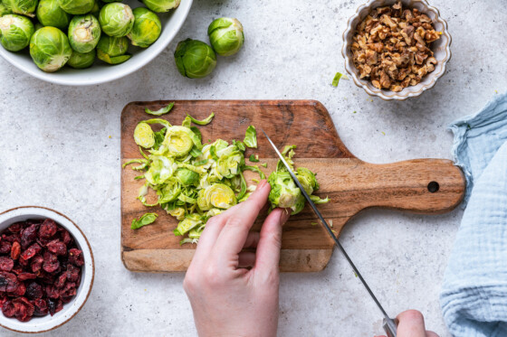 A woman's hand uses a knife to chop Brussels sprouts on a wooden cutting board.