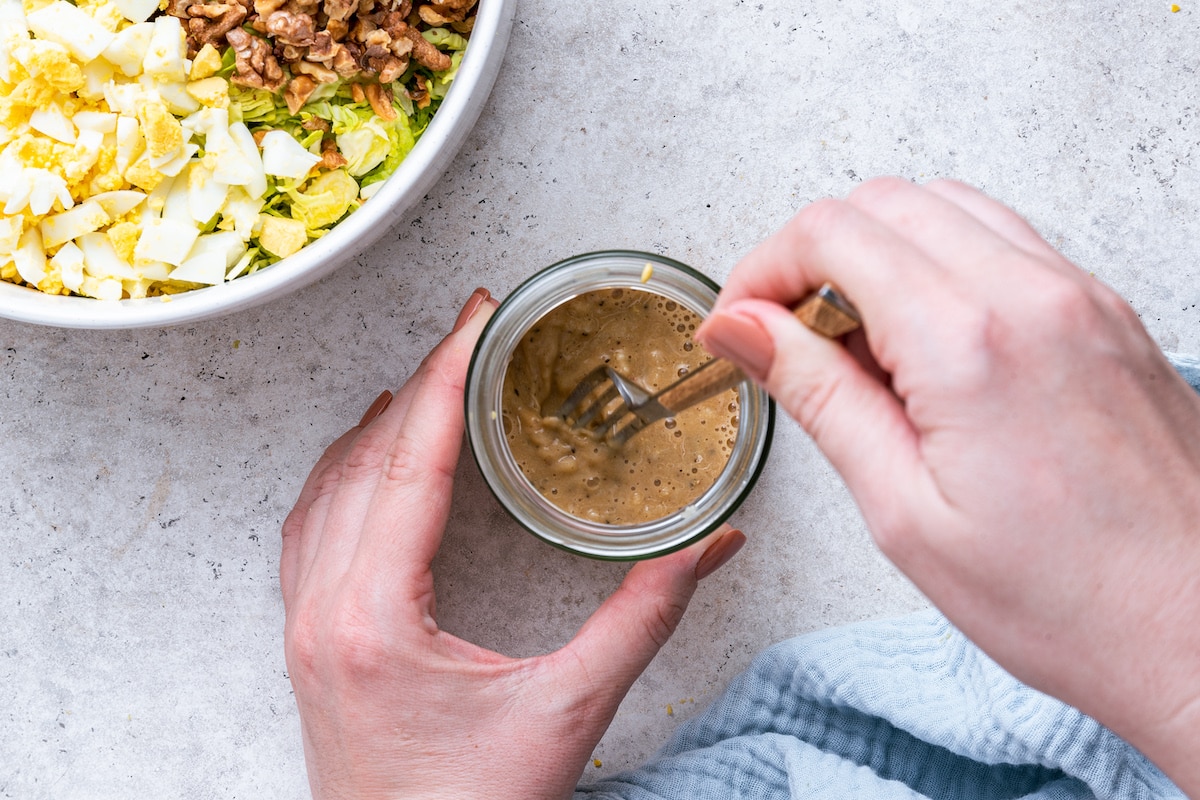 A woman's hand using a fork to mix the ACV dressing in a small glass jar.