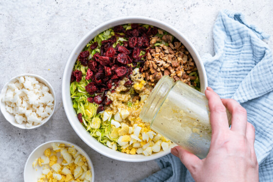 A woman's hand pouring an ACV dressing into a bowl of separated ingredients used to make the Brussels sprout chopped salad. Ingredients include Brussels sprouts, hard boiled eggs, dried cranberries, chopped walnuts, and crumbled goat cheese.