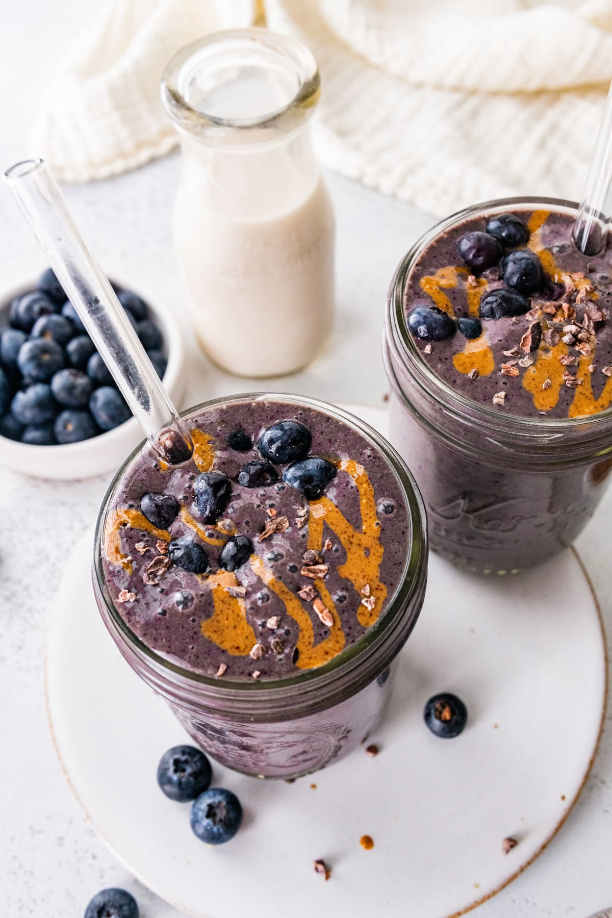 Two chocolate blueberry smoothies with glass straws topped off with fresh blueberries and a drizzle of almond butter.