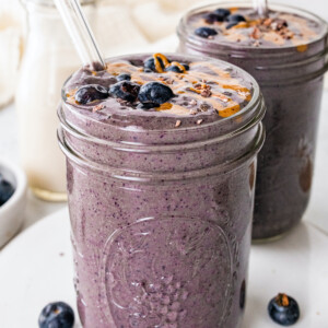 Two chocolate blueberry smoothies with glass straws topped off with fresh blueberries and a drizzle of almond butter.