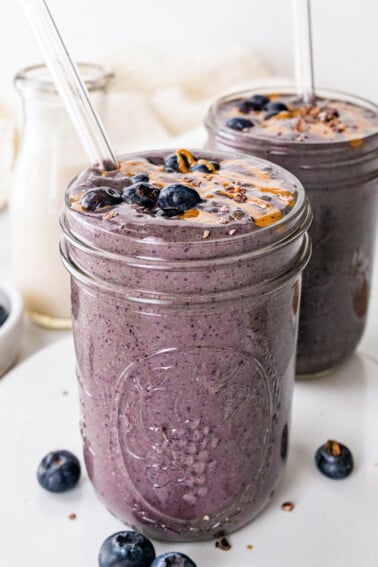 Two chocolate blueberry smoothies with glass straws topped off with fresh blueberries and a drizzle of almond butter.