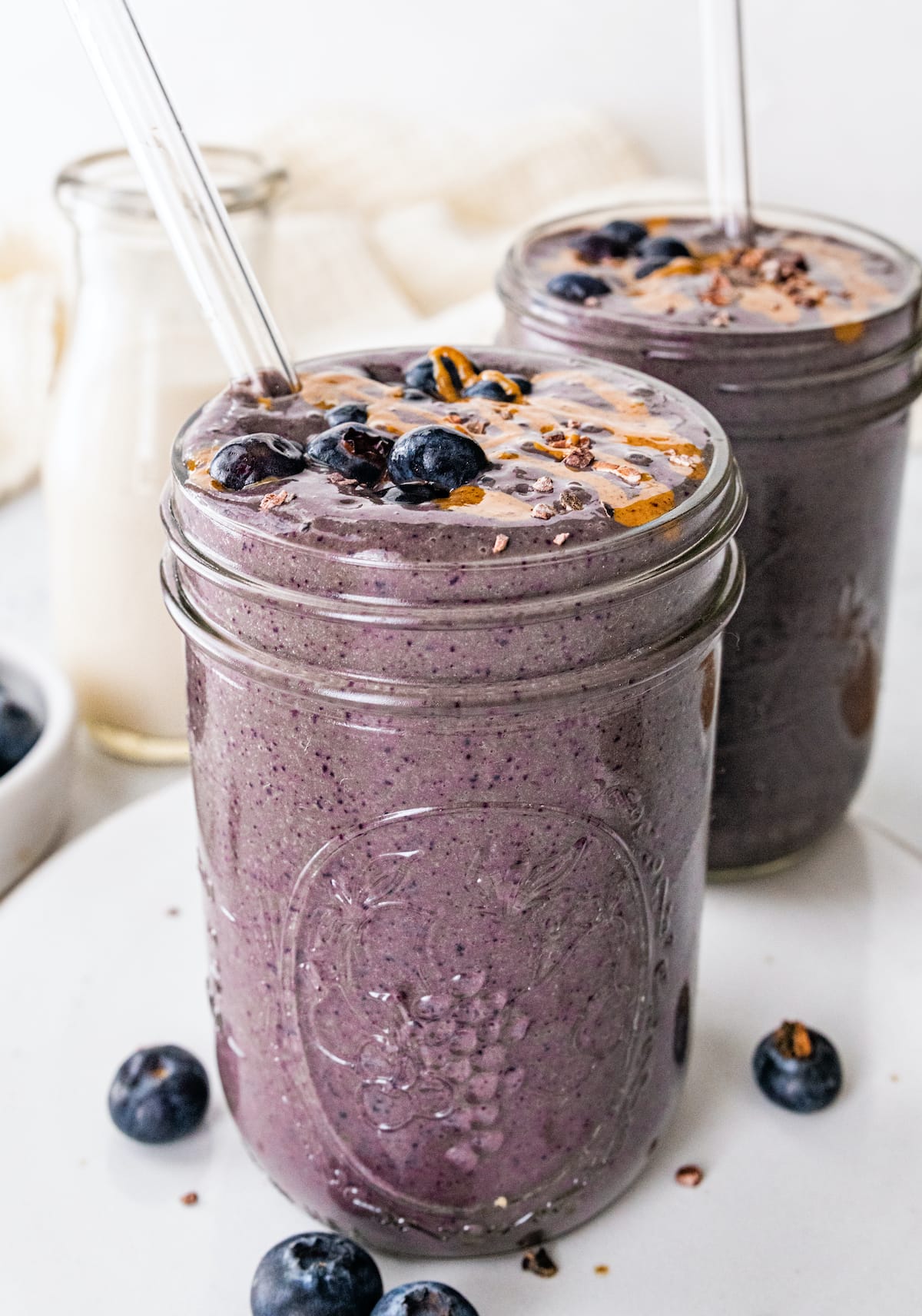 Two chocolate blueberry smoothies with glass straws topped off with fresh blueberries and a drizzle of almond butter.