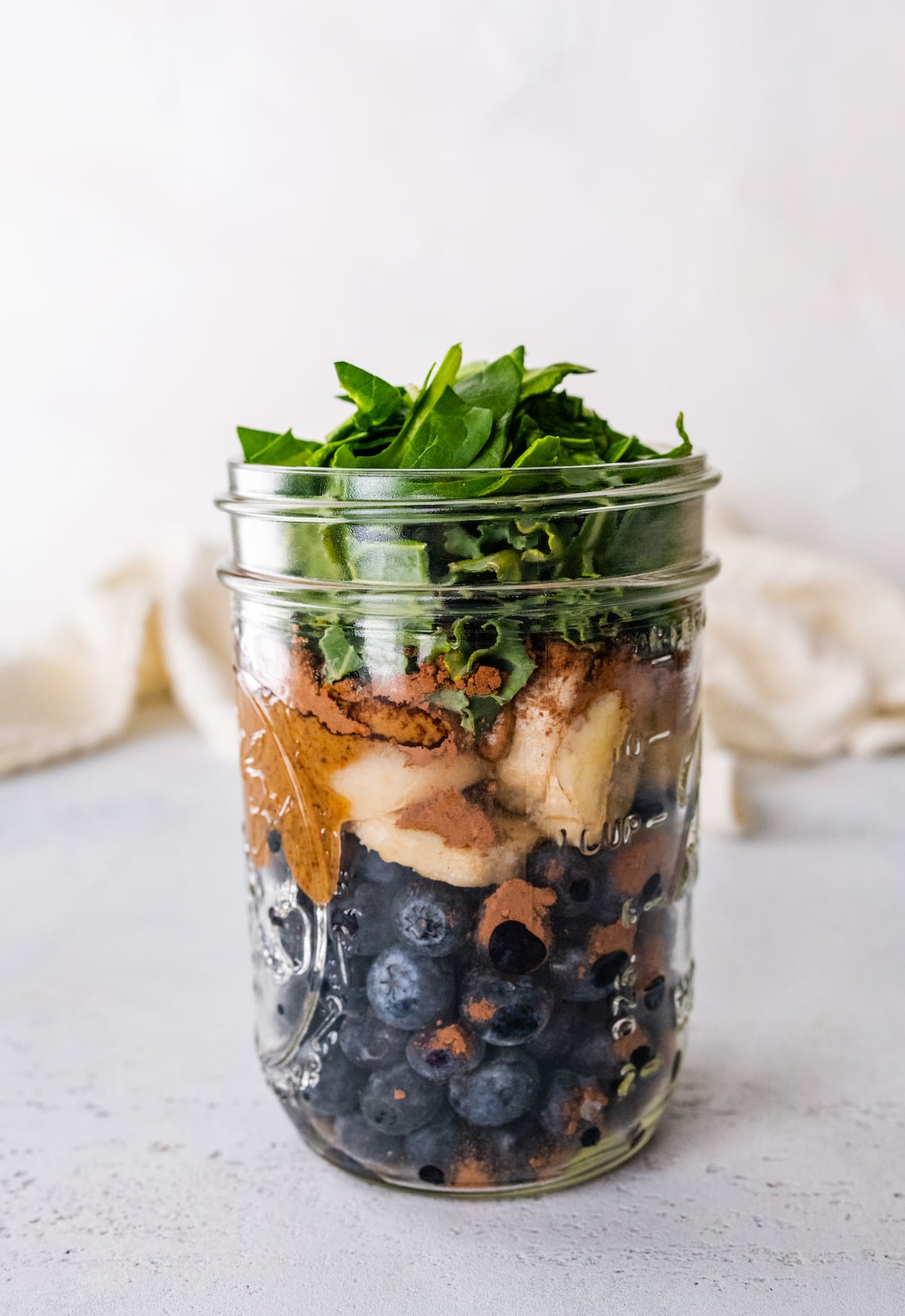 A mason jar with the ingredients for the chocolate blueberry smoothie before being blended. Noticeable ingredients are blueberries, banana, almond butter, cacao powder, and spinach.