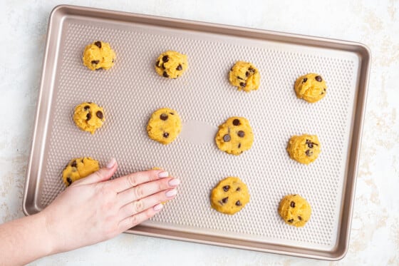 Twelve coconut flour chocolate chip cookies on a baking tray before being baked. A woman's hand is pressing one down to make a more uniformed shaped cookie.