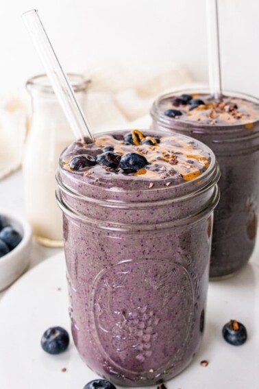 Two chocolate blueberry smoothies with glass straws topped off with fresh blueberries and a drizzle of almond butter.