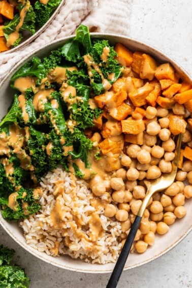 A white bowl containing kale, sweet potato, chickpeas, brown rice, and a creamy peanut sauce.