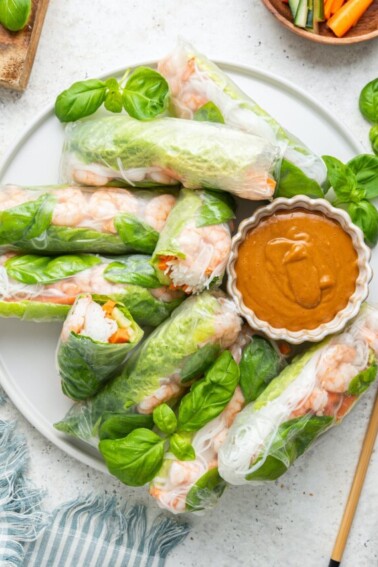 A large white plate containing multiple shrimp spring rolls, some cut in half and a peanut sauce in a small white bowl. Wooden chopsticks are near the plate, and fresh basil leaves are used as a garnish.
