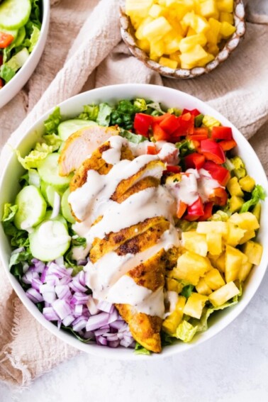 A white bowl containing cooked sliced chicken on top of a bed of lettuce, red onion, cucumber, red bell pepper, pineapple, and a creamy dijon mustard dressing.