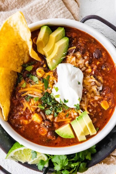 A white bowl full of turkey chili, topped sour cream, shredded cheese, avocado slices, fresh cilantro, and tortilla chips.