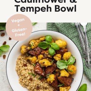 A white bowl containing the curried cauliflower and tempeh over a bed of quinoa and garnished with fresh basil leaves.