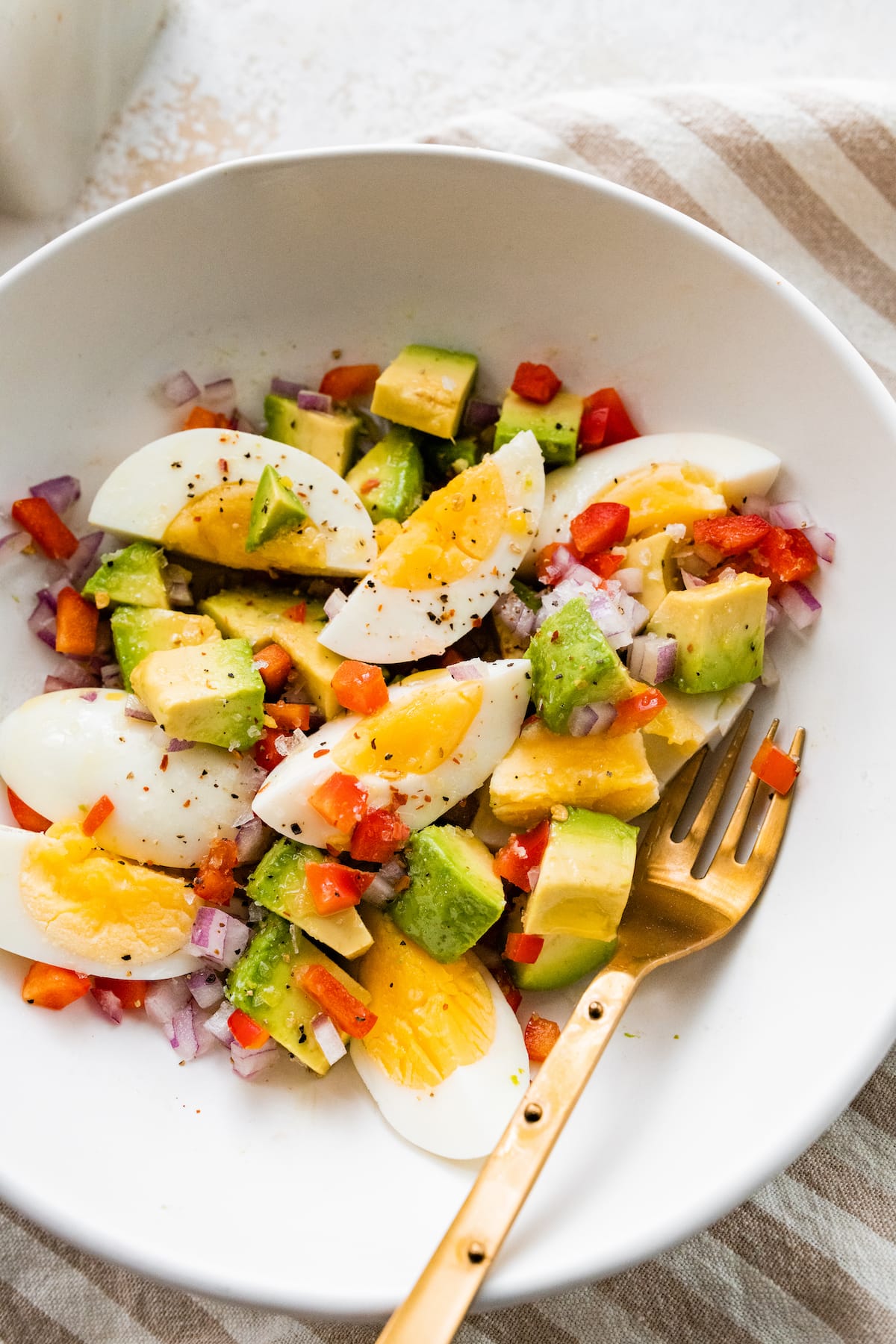 A white bowl with a golden fork containing hard boiled eggs, avocado, red onion, red bell pepper, and topped with spices.