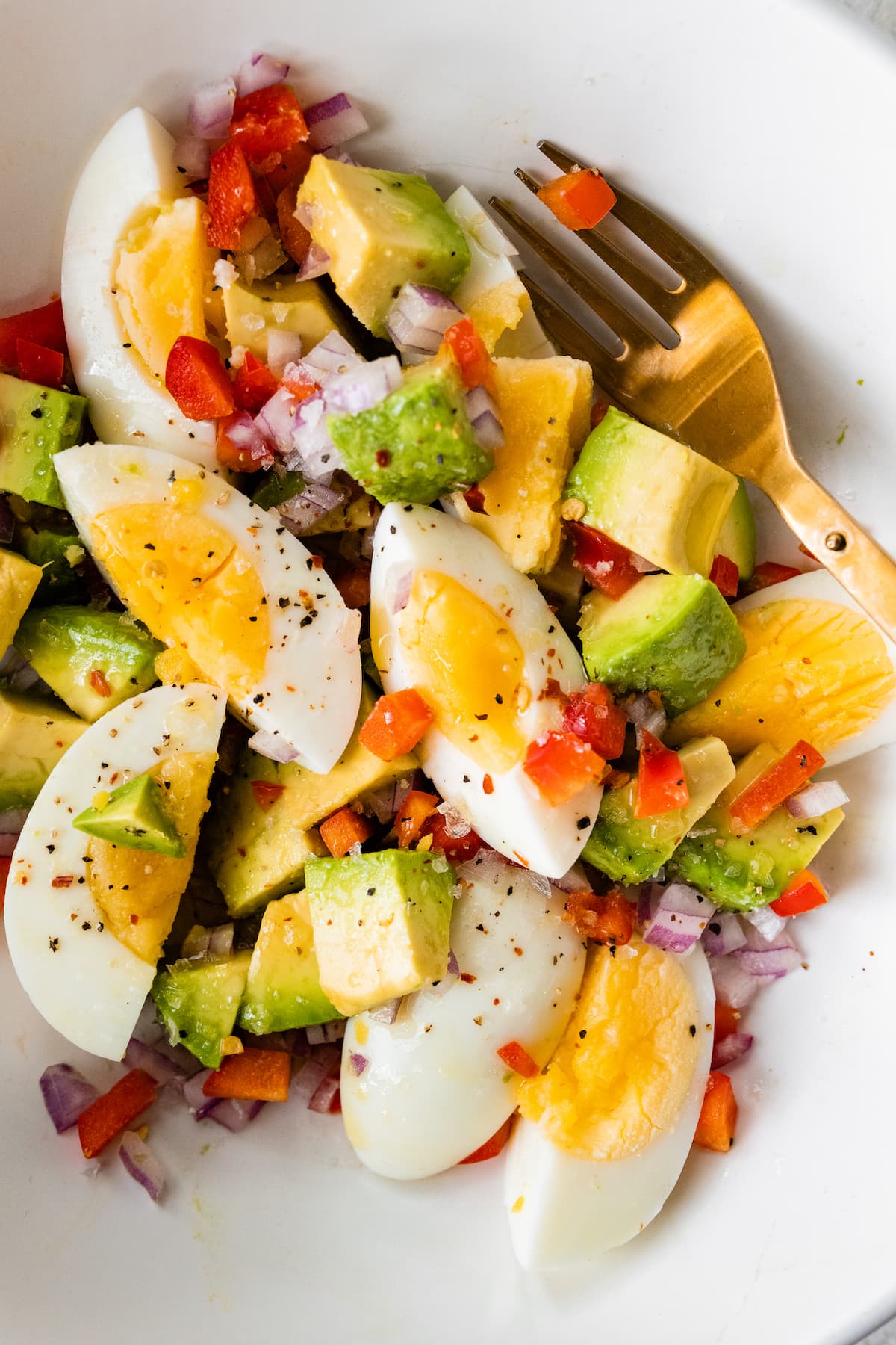 A white bowl with a golden fork containing hard boiled eggs, avocado, red onion, red bell pepper, and topped with spices.