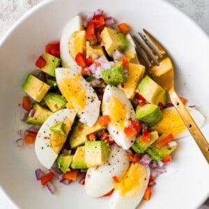 A white bowl with a golden fork containing hard boiled eggs, avocado, red onion, red bell pepper, and topped with spices.