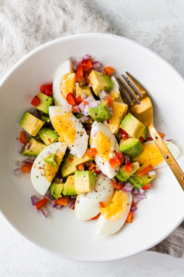 A white bowl with a golden fork containing hard boiled eggs, avocado, red onion, red bell pepper, and topped with spices.