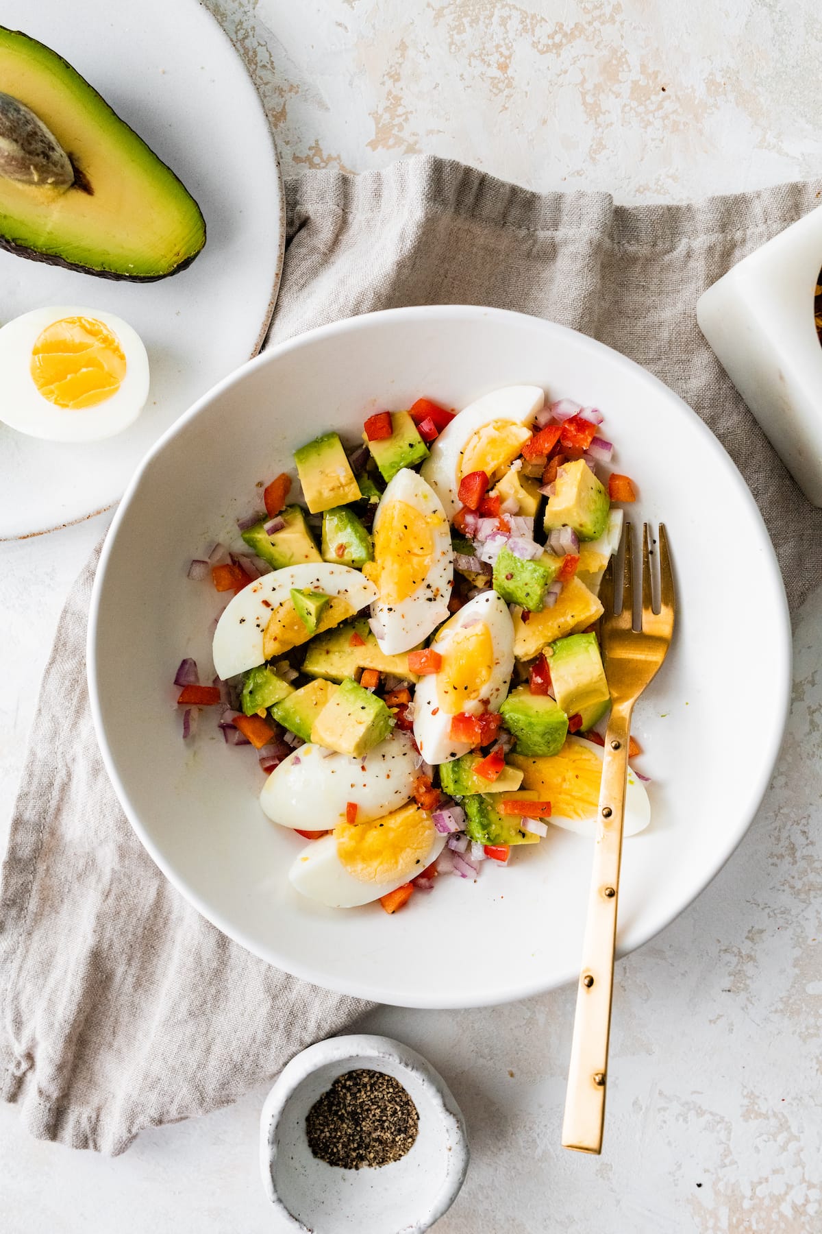 A white bowl with a golden fork containing hard boiled eggs, avocado, red onion, red bell pepper, and topped with spices.