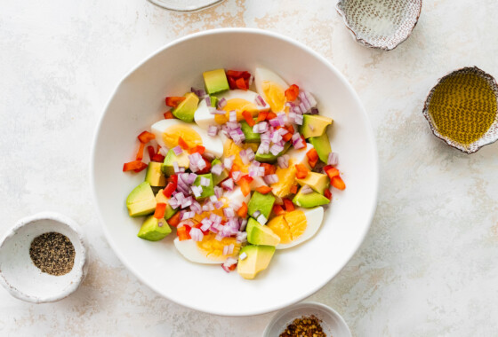 A white bowl containing hard boiled eggs, avocado, red onion, and red bell pepper.