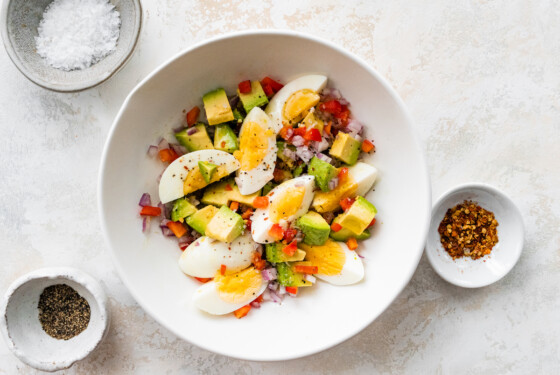 A white bowl containing hard boiled eggs, avocado, red onion, red bell pepper, and topped with spices.