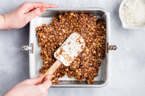 A woman's hand using a silicone spatula to spread the no-bake chocolate coconut bar mixture in a square baking pan.