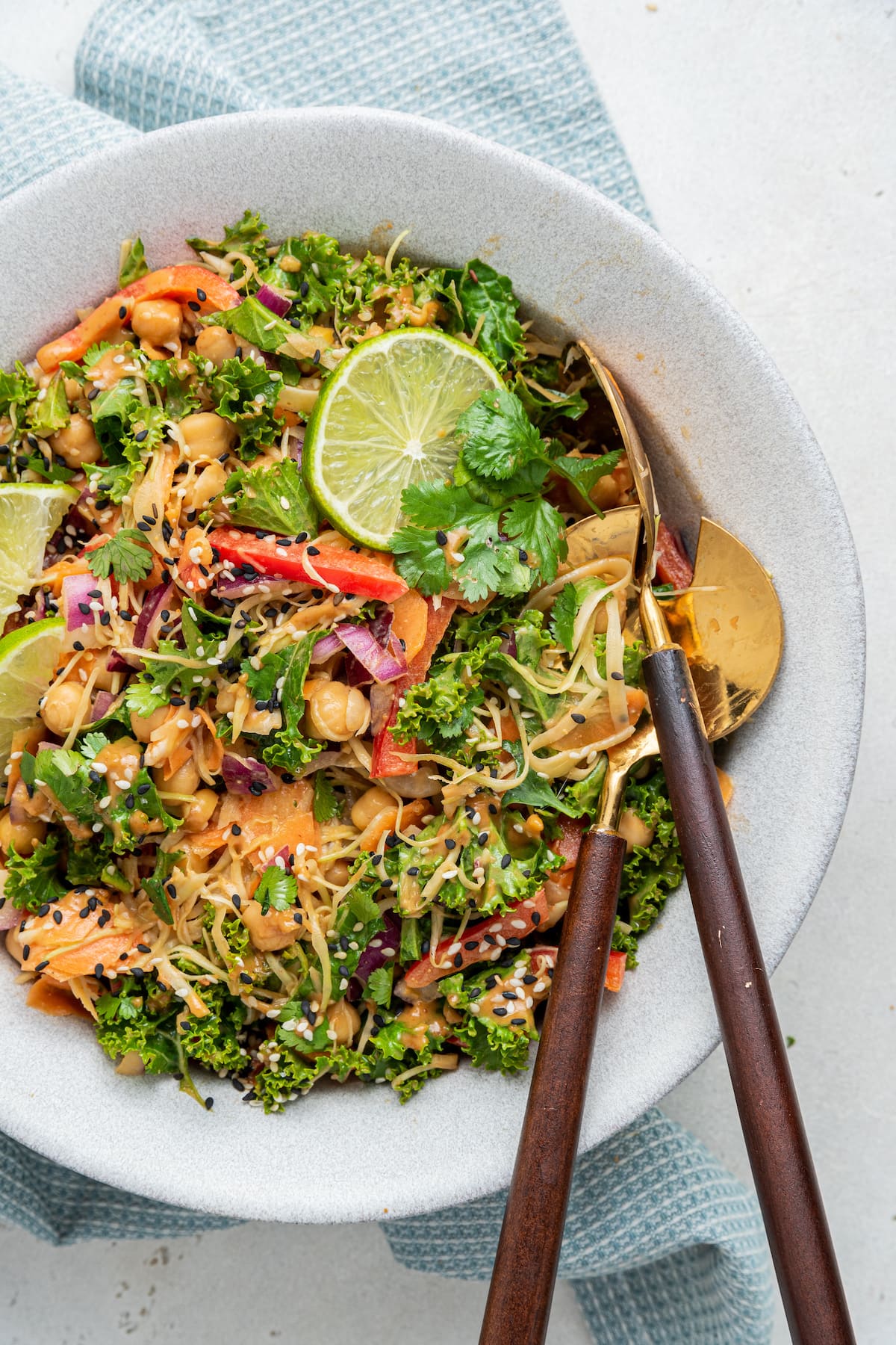 A white bowl with the kale and cabbage pad thai salad, two serving spoons, and a few lime wedges on top.