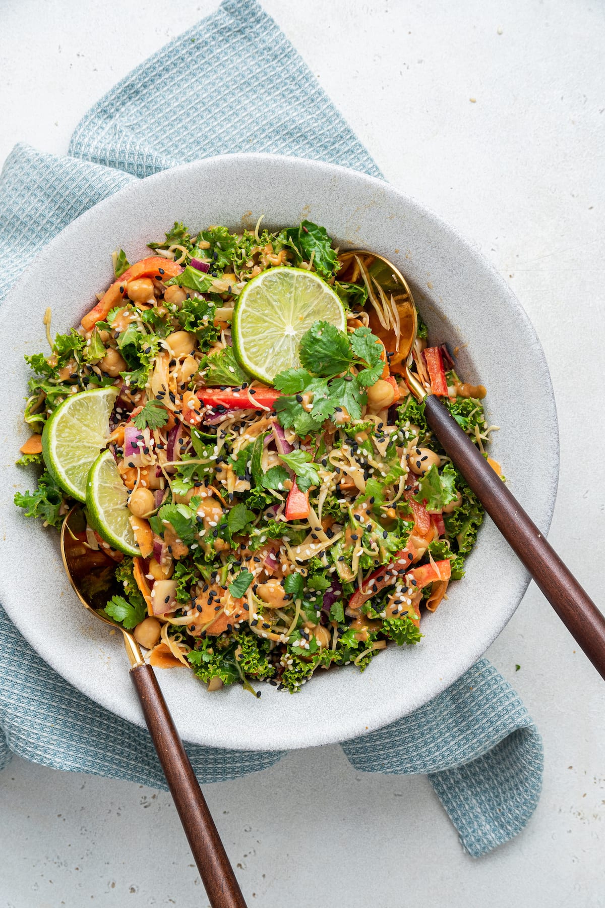 A white bowl with the kale and cabbage pad thai salad, two serving spoons, and a few lime wedges on top.