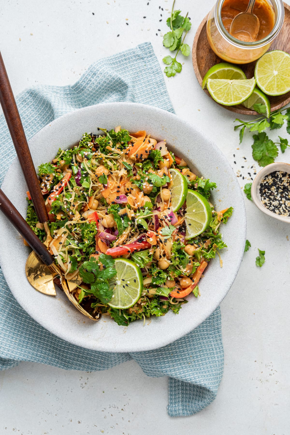 A white bowl with the kale and cabbage pad thai salad, two serving spoons, and a few lime wedges on top.