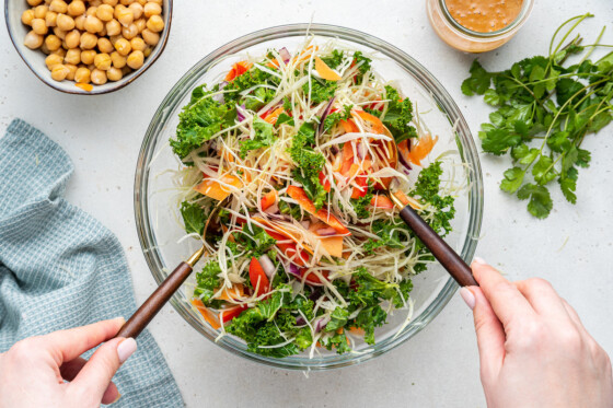 Woman's hands using two utensils to mix the kale and cabbage salad in a large glass bowl.