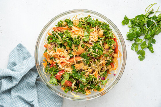 A large glass bowl full of fresh salad. Ingredients include kale, cabbage, red bell pepper, red onion, carrots, cilantro, and chickpeas.