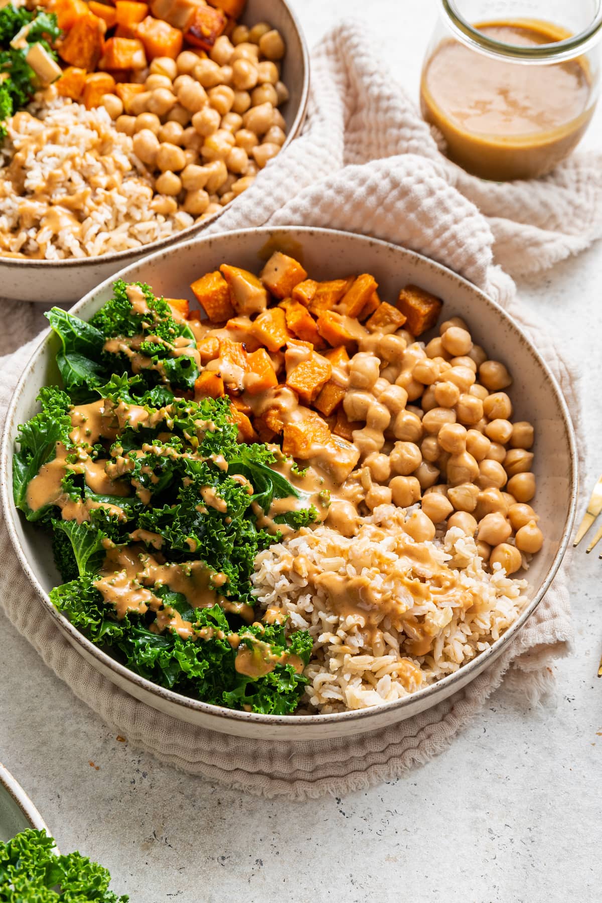 A white bowl containing kale, sweet potato, chickpeas, brown rice, and a creamy peanut sauce.