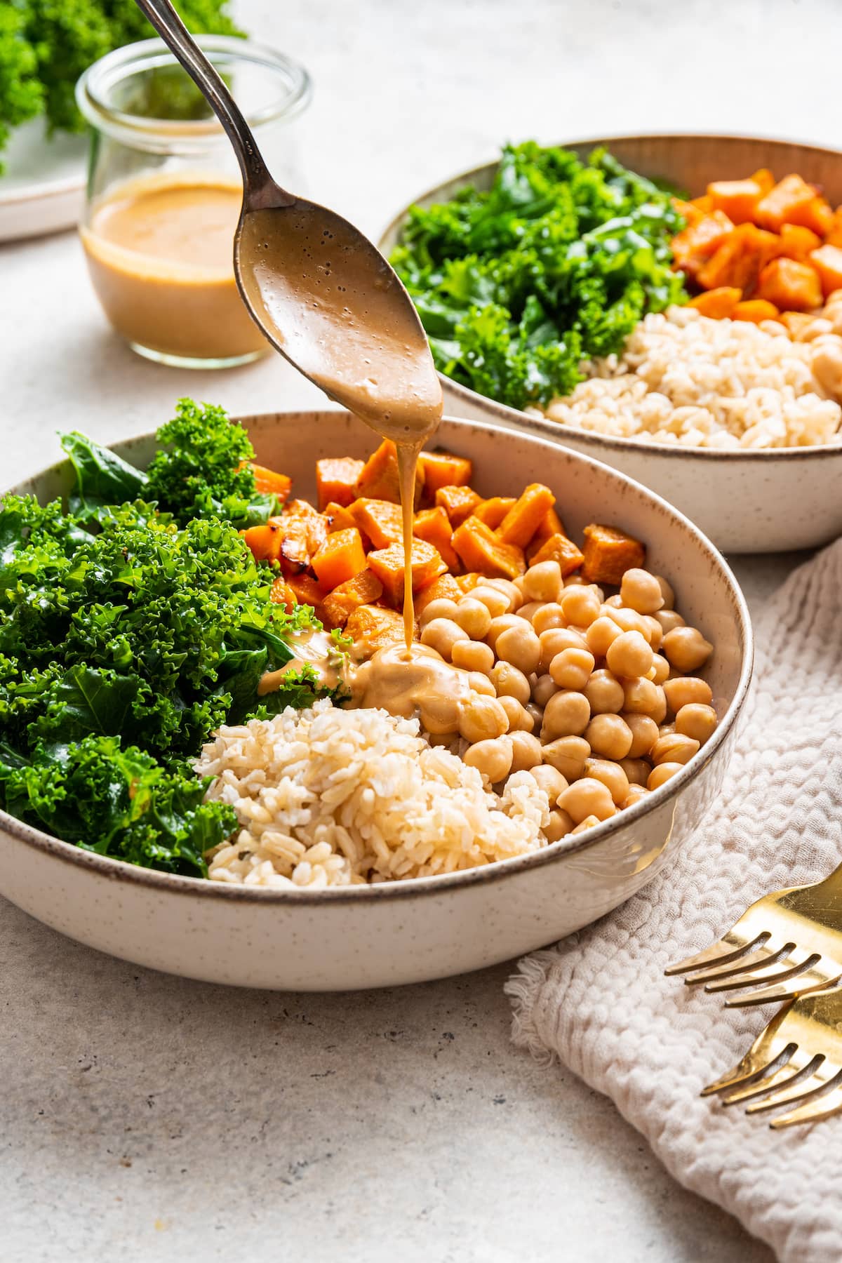 A spoon drizzles peanut sauce into a bowl of chickpeas, kale, brown rice, and sweet potato.