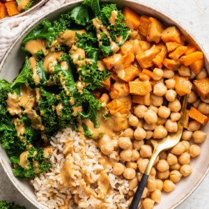 A white bowl containing kale, sweet potato, chickpeas, brown rice, and a creamy peanut sauce.