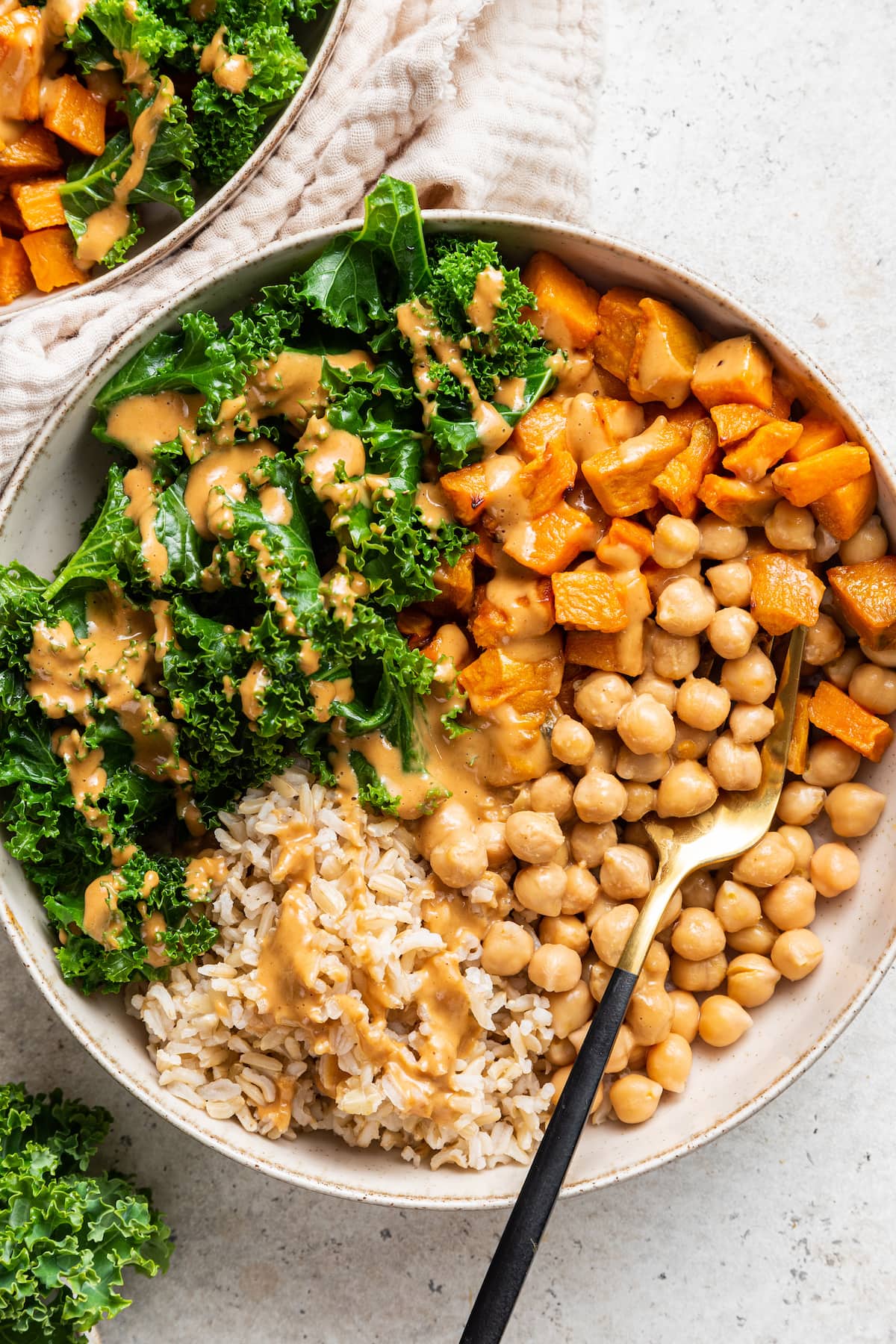 A white bowl containing kale, sweet potato, chickpeas, brown rice, and a creamy peanut sauce.