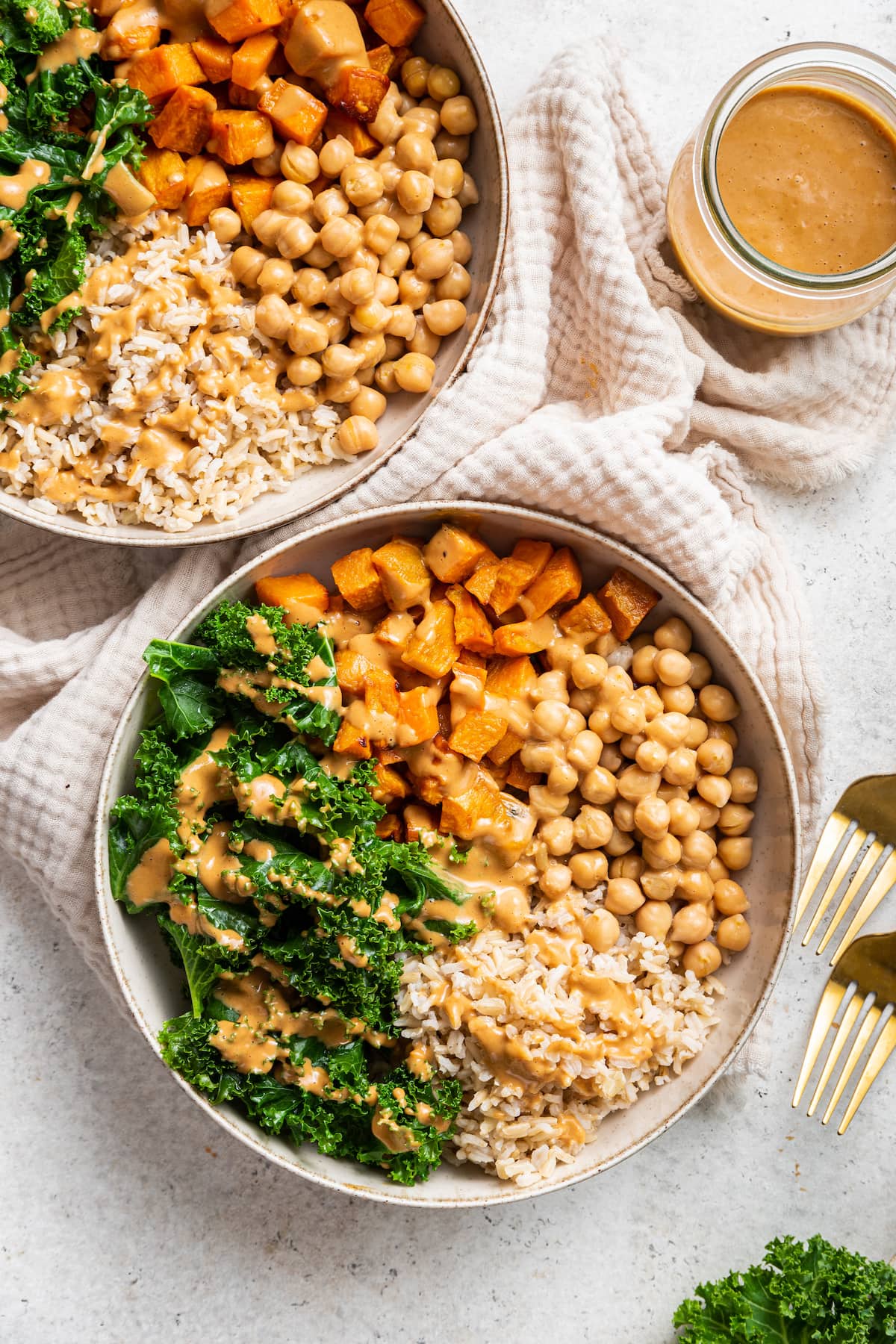 Two white bowls containing kale, sweet potato, chickpeas, brown rice, and a creamy peanut sauce.
