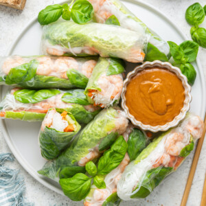 A large white plate containing multiple shrimp spring rolls, some cut in half and a peanut sauce in a small white bowl. Wooden chopsticks are near the plate, and fresh basil leaves are used as a garnish.