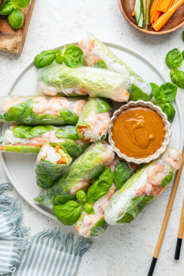 A large white plate containing multiple shrimp spring rolls, some cut in half and a peanut sauce in a small white bowl. Wooden chopsticks are near the plate, and fresh basil leaves are used as a garnish.