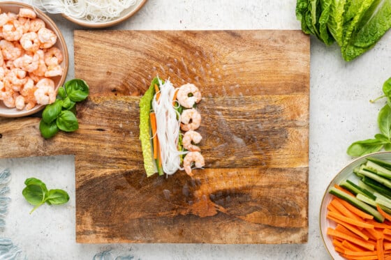 Shrimp, lettuce, carrots, vermicelli noodles, and cucumber on top of a wet rice paper wrap on a wooden cutting board.